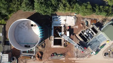 An aerial view of a storage tank. The very large cylindrical structure on the left of the patch of land has blue pipes coming off it. There is a rectangular hole and work equipment, including a white crane, dotted around the rest of the site.