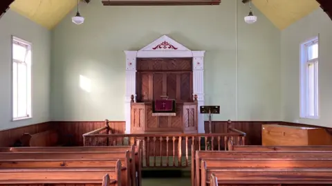 View of the inside of the chapel. Pews on either side with an ornate pulpit in the centre on the far wall. There's a piano or organ on the far right side. 