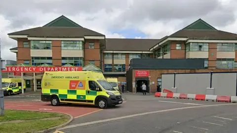 The Accident and Emergency department at the Royal Bolton Hospital. There are ambulances parked outside the building and one appears to be driving away. The ambulances are bright fluorescent yellow with green checks.