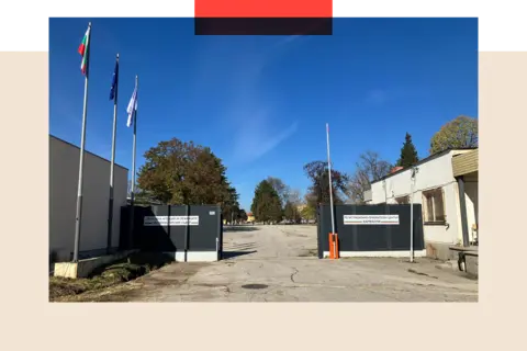 Photo of the exterior of the Harmanli exile  camp. The main   gross  is unfastened  with the car   obstruction  raised and a factual  expanse behind. There are trees and the entity  is blue.