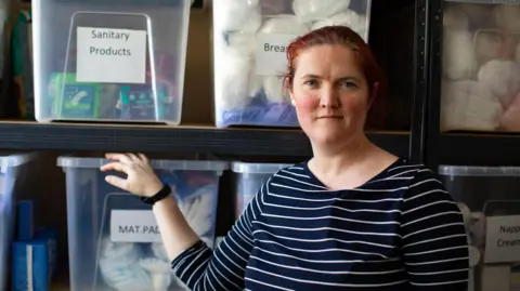 Emilie de Bruijn is in her early 40s with dark red hair pulled back into a pony tail. She is wearing a black and white top with stripes and is standing in front of a load of boxes containing children's clothing. 