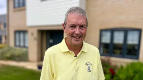 A man wears a yellow T-shirt with a logo on his left hand side. He is standing in front of a modern house with a front garden. 