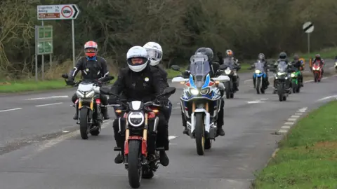 Andy Simons Line of motorbikes passing a sign to Cogenhoe