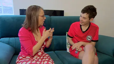 A woman wearing a pink spotted dress and a plain pink cardigan sits on the left of a soft, teal sofa. She is facing her teenage son, sat on the right of the sofa in a bright pink Brighton and Hove Albion kit.