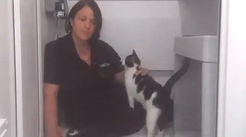Vickie Blackburn strokes black and white rescue cat Hattie. She is sat on the floor wearing a black uniform.
