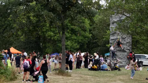 A large group of people, mostly women with children, gathered around in a park which is hosting a summer event. There are two stalls with awnings, set among the trees, and a high mobile climbing wall with two people starting to ascend.