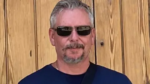 Dean Young Dean Young wearing dark glasses and a dark blue T-shirt and standing against a wooden door. He has greying hair and a greying goatee beard and is starting to smile. 