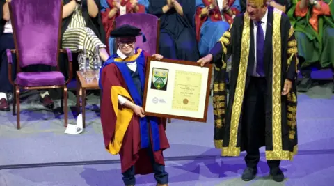 Leeds Beckett University A picture of George Webster in his graduation gowns being presented with a framed honorary degree certificate. There are people sitting behind him.
