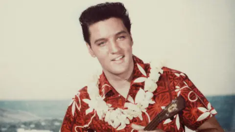 Getty Images American rock n' roll singer Elvis Presley plays a ukelele, wearing a Hawaiian shirt and lei, in a still from the film 'Blue Hawaii'.