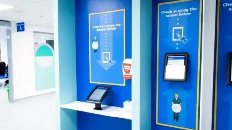 Cambridge University Hospitals NHS Foundation Trust The inside of a hospital with a sign that reads: "Check in using the screen below" with an arrowing pointing down towards an ipad. 