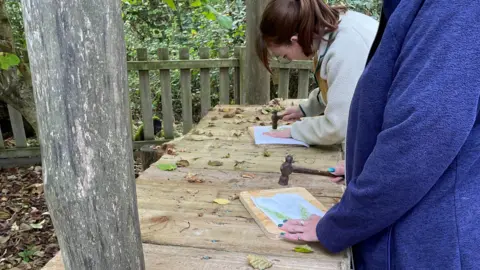 Alex Lister Two people lean over a wooden desk at the woodland classroom and use wooden hammers to work on a craft project using paper, leaves and wooden boards.