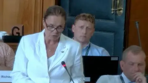 Devon County Council Reform UK councillor Sue Davies in a white jacket over a white top and wearing glasses, she is speaking into a microphone in the Devon County Council chamber