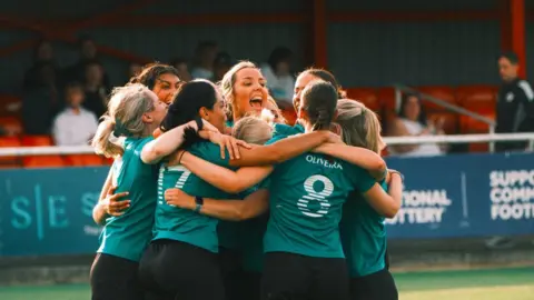 Aston Martin F1/XA Group de mujeres vestidas con un kit de fútbol verde y negro se abrazan en un grupo y celebran en un campo de fútbol. Detrás de ellos hay un estadio con miembros del público que los observan.