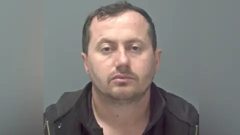 A police custody photo of Jani Murati, a man with short dark hair staring straight into the camera