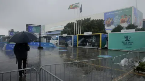 Reuters A man stands outside the Palacio de Deportes Jose Maria Martin Carpena Arena in Malaga holding a navy umbrella while torrential rain surrounds him. The man is looking at the tennis arena as a number of flags, including the Spanish and Andalusian flags, flutter in the wind