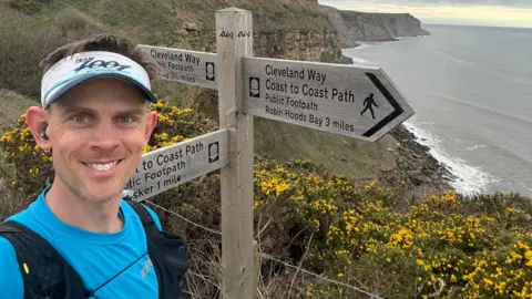 Ben Morris Runner Ben Morris stands in front of a wooden sign which has directions to the Cleveland Way and coast-to-coast path. He has short brown hair and is wearing earphones, a blue t-shirt and a black running rucksack. The sign is on the side of a cliff which has yellow hawthorn and the sea in the background. 