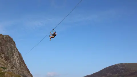 A person suspended from a zip line travels high above mountains, centered against a wide, clear blue sky. Rocky slopes rise on the left and right edges of the image.