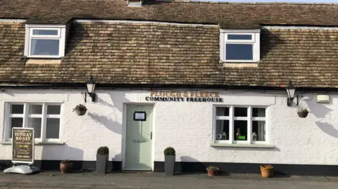 Bev Rimmer/BBC The white-brick front of a public house. It says "PLOUGH AND FLEECE" in-between a green front door and a white window. The lettering is in capitals, and is gold-coloured. Underneath this, it says "COMMUNITY FREEHOUSE" in black capital lettering. A black billboard sign to the left of the green door reads: "Traditional Sunday Roast."