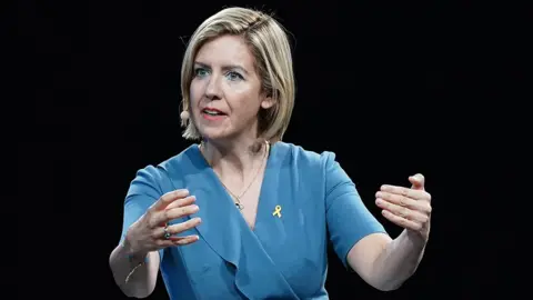 Dame Andrea Jenkyns giving a speech at a conference she has short blond hair and is wearing a blue dress and a headset microphone