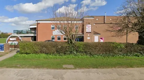 Google A two‑storey brick building with signs for The Sports Bar and Stevens Decorators, partly obscured by a hedge and a tree, with a gated entrance to Dunnington Bowling Club on the left and a grassy area in the foreground under a bright, partly cloudy sky.