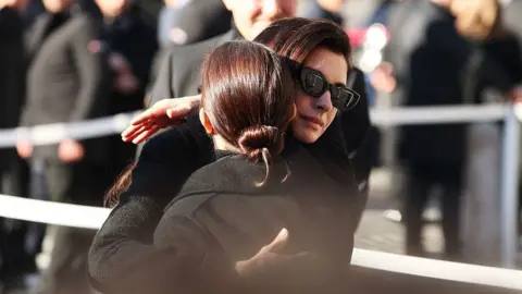 Deepixel/Getty Images Two women dressed in black, one with black sunglasses, embrace outside the church among other mourners