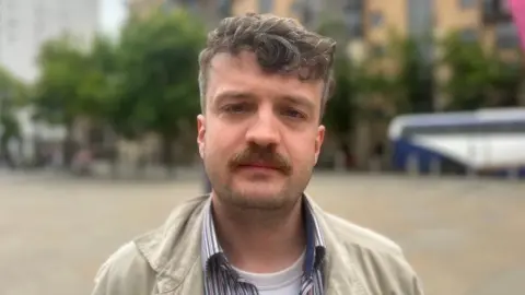 Nathan Young has dark hair and a moustache and stubble. He is wearing a tan jacket, striped open-necked shirt and white t-shirt.