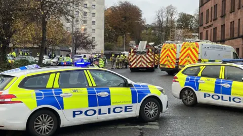 BBC Two police cars in front of shot, with fire engines and fire crews behind and the bottom floors of the tower block also visible in background