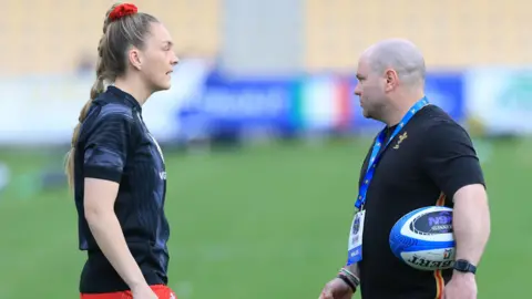 Hannah Jones in a training top talks to Sean Lynn who has a rugby ball under his arm on a pitch before a game.