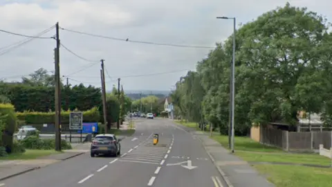 London Road in Benfleet. A single car is visible, driving away form the camera. There are bushes, front gardens and houses on the righthand side