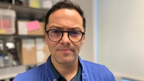 Prof Alvaro Mata wearing round, black-framed glasses looking down the camera. He has a blue overshirt on.