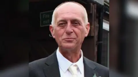 Kent Police Alan Tomkins looks at the camera and is smiling. He is wearing a dark suit with a white tie and white shirt. 