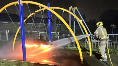 A firefighter putting out a fire on a swing, part of play equipment in a park. His hose, with water coming out, is aimed at the swing. The metal frame is blue and yellow. The image is taken in the dark and buildings are in the distance. 