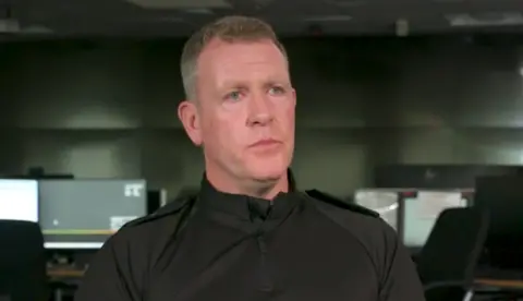 Police Scotland Insp Scott Casey wears a black police uniform and sits in a room with rows of desks with computers behind him. He has short grey hair.