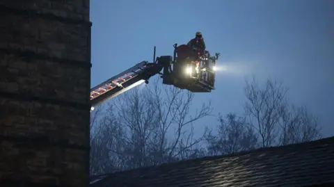 Phil Taylor Firefighter on an aerial platform as he tackles a fire. He is in the air above a roof in the early hours of the morning 