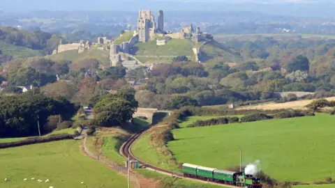 Andrew P.M. Wright/Swanage Railway/PA Wire Corfe Castle photographed from far away on a sunny day. A Victorian steam train can be seen passing through the foreground