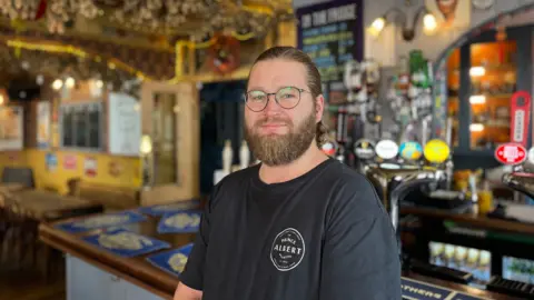 A man with a beard stood in front of a pub bar.