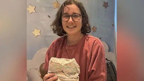 Elizabeth Kane with brown bobbed hair and glasses wears a dark orange top holding a beige and brown patterned reusable nappy. She is smiling. She is in a nursery with a baby behind her.