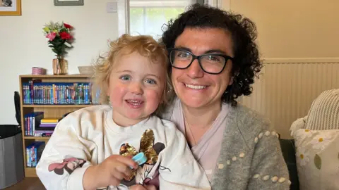 Georgie Murray-Gourlay is holding her daughter Lilly in her arms. Georgie has dark curly hair and Lilly has blonde curls. They are both smiling. They are sitting at home on a sofa, with a bookcase in the background.