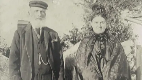 The Kelly Family Collection Black and white photo of an older couple standing in front of a stone wall. He is has a grey beard and it wearing a cap and has a pocket watch chain hanging across his waistcoat, and she has her hair tied up and it wearing a shawl. 