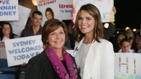 Nancy Guthrie, the 84-year-old mother of US. journalist and television host Savannah Guthrie, who went missing from her home in Tucson, poses with her daughter in front of people carrying signs welcoming them to Sydney, Australia in 2015. 