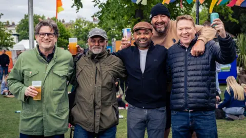 @Slow Moving Media Five men smile at the camera while holding drinks as they stand in an open air part of the Cheltenham Jazz Festival