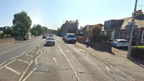 Road with cars and a van on it with houses down either side. There is a pedestrian walking on the pavement.