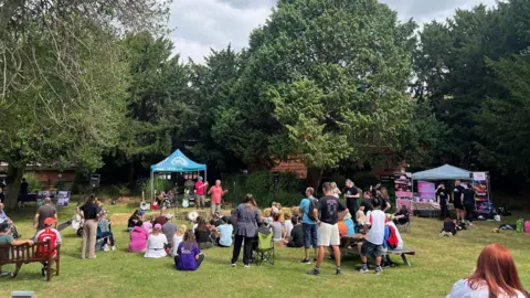 Bedford Inclusive Performing Arts Festival A large group of people outside in a garden watching a performance, with gazebos in the garden. Some are sitting down, others are standing up. There is grass and trees in the distance. 