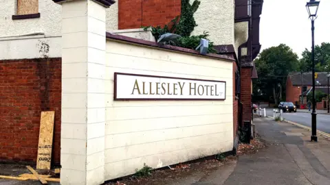 A cream and maroon sign saying "Allesley Hotel" is on a wall outside the premises. A road runs alongside.