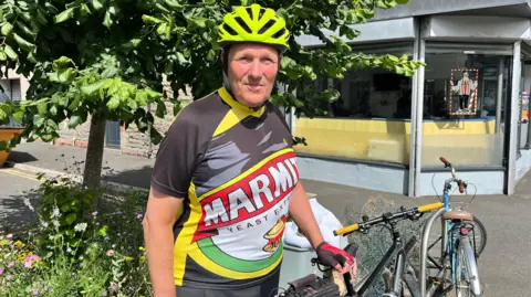 Patrick stood next to his bike on Cathays Terrace. He's wearing a neon green helmet with a Marmite cycling shirt on, and is stood outside a barbers and beside a tree. 