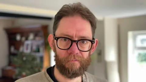 A head and shoulders shot of a man looking directly into the camera with short ginger hair and a ginger beard. He is wearing black-framed glasses and has a worried expression. A window and bookshelf can be seen in the background. 