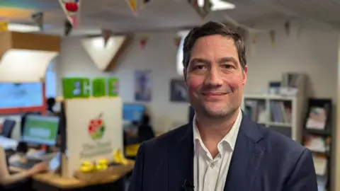 Nathan Standley / BBC Myles Bremner smiles into the camera in a head and shoulders shot. He is wearing a navy suit jacket over an open-collared white shirt. He stands in front of a blurred office background with staff working on desktop computers.