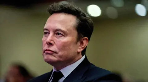 Reuters Elon Musk memalingkan muka dari kamera. Ia mengenakan jas berwarna gelap, kemeja putih, dan dasi berwarna gelap. 