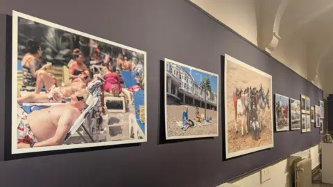 Jadzia Samuel/BBC Some of the art provided for JJ Waller's Beside the Sea exhibition at the Hastings Museum and Art Gallery. Several images, primarily of people sunbathing at the beach, can be seen. All pictures are set against a purple wall.
