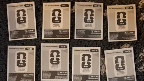 Eight grey stickers sit on a darker grey rug. They are the back side of Panini 2026 World Cup stickers They read QAT5, QAT11, GER14, EGY16, FRA16, AUT6, ENG3, PAN3. The Fifa 2026 World Cup logo an Panini logo are visible. 